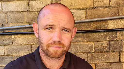 A bald man with a short beard, wearing a black shirt, gives a slight smile to the camera.