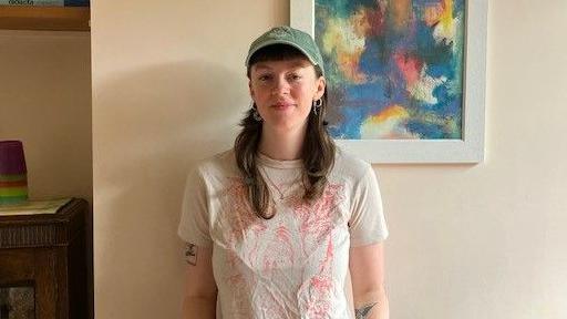 A woman standing against a cream wall. She is wearing a white top and has tattoos on her arms. She is smiling and wearing a baseball cap.
