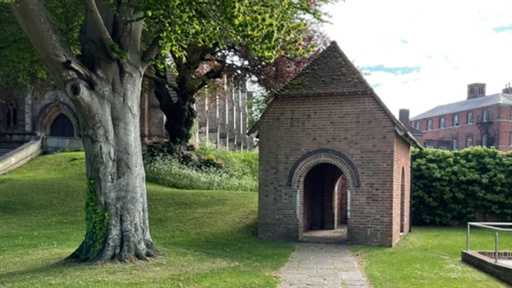 A red brick outdoor shelter, with pitched roof and arched entrances on three sides, on a green lawn next to a large tree with a large chapel behind.