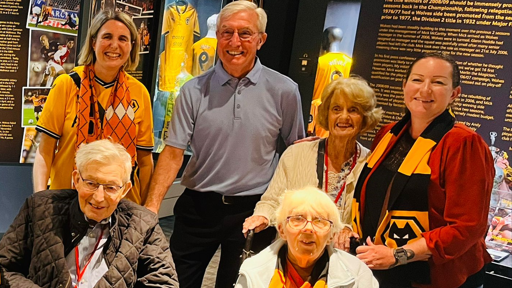 Three of the residents pose for photos with John Richards, who has white hair and is wearing a blue polo shirt and black trousers with a belt. Two of the residents, a man and woman, both with white hair, are seated. The other resident, a woman with curly white hair, is standing behind them. Two other women, one with bobbed blonde hair and one with dark hair tied back, are standing either side and wearing Wolves scarves and kit. Behind them you can see club images and memorabilia.