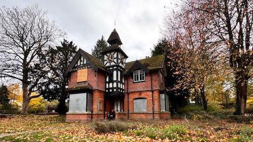 A building stands in a park. It is made of brick and has sections in black and white with a clock tower and a turret. Most of the windows are boarded up.