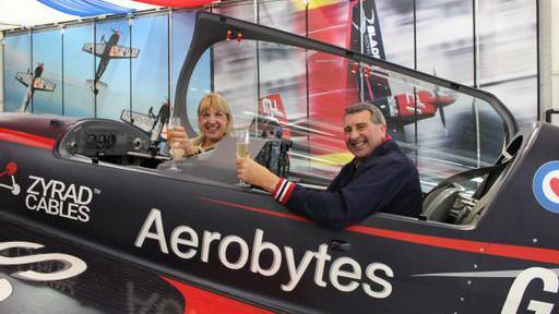 David Traynor and Angie sitting inside an aircraft holding glasses of champagne.