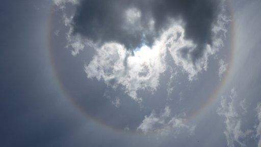 A circular, rainbow coloured band of light surrounds the sun which is covered by cloud