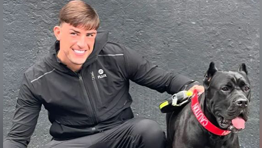 Jack Fincham crouched on the ground next to a black cane corse dog. He is wearing a black tracksuit and is smiling at the camera. The dog wears a red collar/lead.