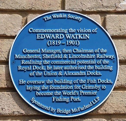 A blue plaque on a redbrick wall. It has white writing that reads "The Watkin Society. Commemorating the vision of EDWARD WATKIN (1819 - 1901) General Manager, then Chairman of the Manchester, Sheffield, Lincolnshire Railway. Realising the commercial potential of the Royal Dock, he later authorised the building of the Union & Alexandra Docks. He oversaw the building of the Fish Docks, laying the foundation for Grimsby to become the World's Premier Fishing Port. Sponsored by Bridge McFarland LLP".