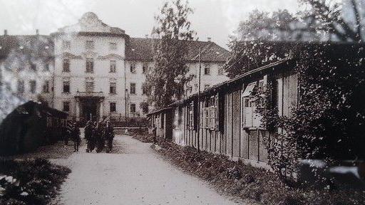 A black and white photograph of a large, old building with multiple windows and a central entrance in the background. A dirt path leads toward the building. On the right side of the path are smaller wooden structures covered in ivy or vegetation. A group of people stands on the left side of the path near the entrance to one of the smaller structures. The scene has a historical and aged appearance