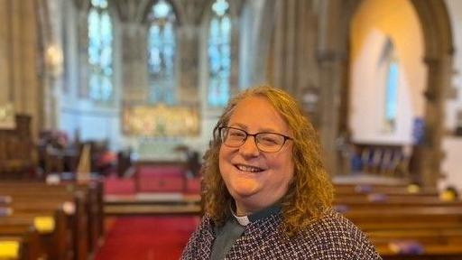 A smiling Reverend Heather Atkinson stands in the middle of her church. The altar and ornate stained glass windows stand behind her. She has curly blonde hair and glasses and is wearing a grey top with white dog collar and a purple-and-white patterned jacket.