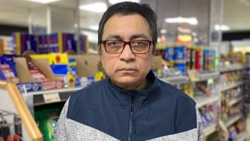 A man wearing a blue and grey zipped jumper with dark hair and glasses is standing in a convenience story with items on the shelves behind him.