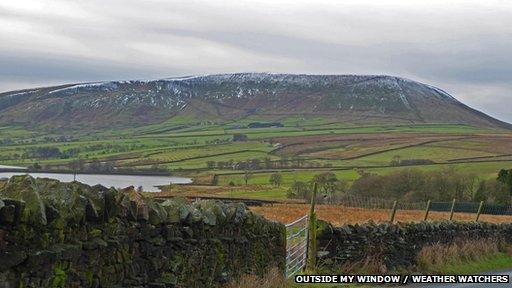 In the distance is a very high hill, with a small amount of snow on it. In the foreground are green fields and a stone wall