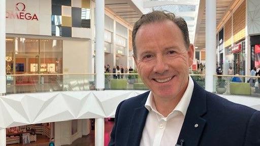 A smiling man with short, brown hair who is wearing a white shirt and blue suit jacket. In the background are several shops and shoppers.