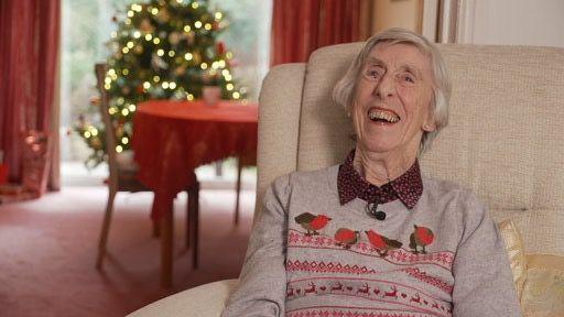 Margaret Lines, wearing a Christmas jumper, smiles as she sits in a chair in what looks like a living room. There is a table with a red tablecloth and a Christmas tree with its lights on in the background. 