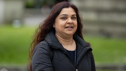 A slightly smiling Tanya Nasir, who has long curly dark hair and is wearing a black jacket and silver chain, looks side on to the camera with a blurred green background behind her