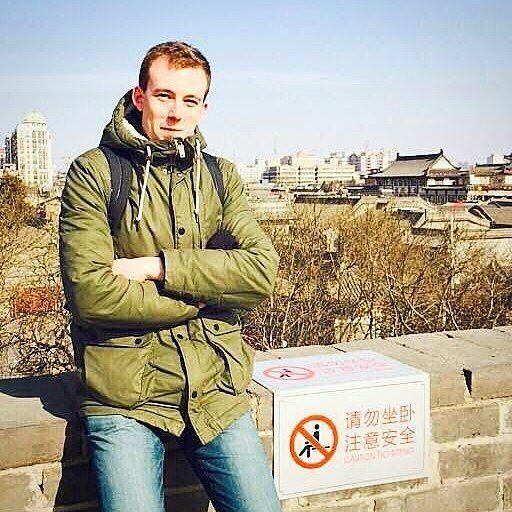 Christopher Berry pictured sitting on a wall in China. He is wearing a green coat and jeans and has a backpack on. Behind him buildings in a Chinese style can be seen and there is a sign with Chinese characters