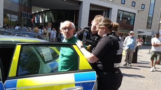 James Grote, in a green shirt and clerical collar, is put into a police car by two officers.