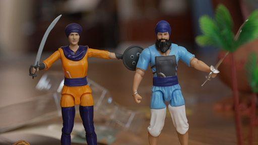 Two toys stand on a table - a female character in orange and purple clothing with a sword and shield and a male character in blue clothing with a sword. Both wear turbans