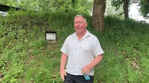 A man with short white and brown hair, stands in front of a grassy bank with a metal plaque halfway up it. He wears a white short-sleeves shirt with a blue dotted pattern while holding a bottle in his left hand.
