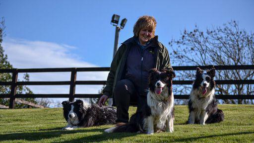 Andrea Bainbridge She is wearing a fleece and a coat and is kneeling next to her three border collie dogs. They are in a grass-covered field with a wooden fence behind them.