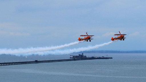 Two red stunt planes flying side by side above the sea in Southend, with a trail of grey smoke behind them. The city's pier is pictured in the background, stretching out to sea.