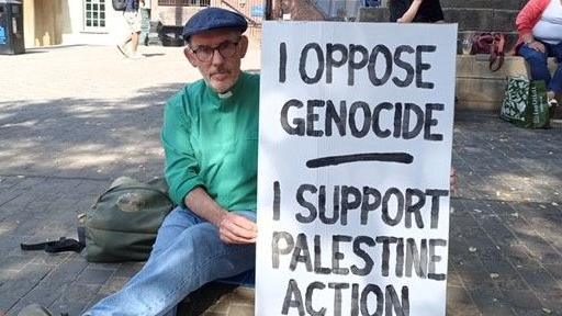 James Grote is wearing a a green shirt, clerical collar and blue hat. He is holding a white placard reading "I oppose genocide, I support Palestine Action".