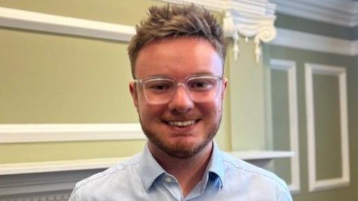 Ted Dolman smiling into the camera. He has brown, short, spiky hair and is wearing glasses. He's also wearing an open collar blue shirt. He's standing in an office within Hull's Guildhall.