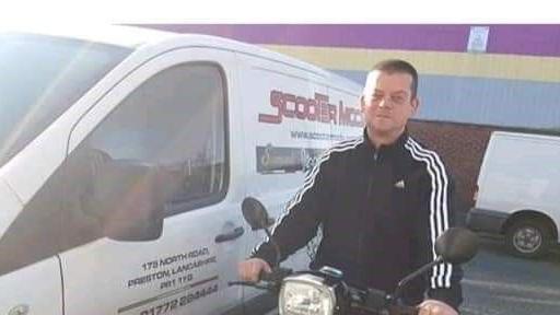 Lee Warburton, with short brown hair, wears a black tracksuit top as he sits on a motorbike parked up near a white van near a warehouse.