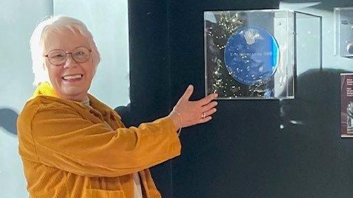 Gail Morecambe wearing a mustard yellow coat and white hair is gesturing to a blue circle plaque in a clear plastic box. She is smiling at the camera. 