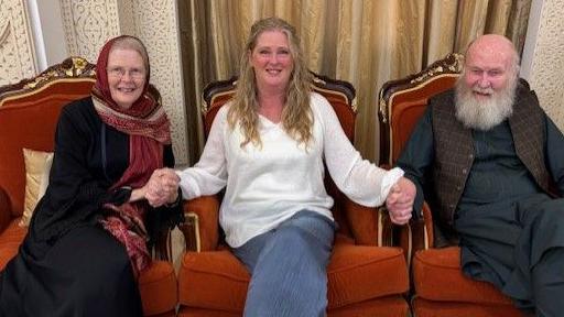 Freed British couple peter and barbie Reynolds sit with their daughter sarah. On the left, Barbie is wearing a dark dress. A blond haired Sarah is sat in the middle of the two, wearing blue jeans and a white top. On the right sits Peter, he's wearing a traditional Afghan dark top and trousers, with a brown textured waistcoat. All are smiling at the camera.