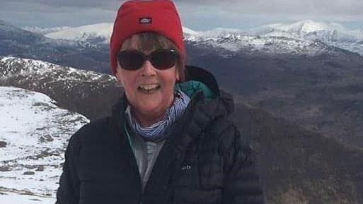 Sally Hall standing on top of a snowy mountainside wearing a red hat and dark sunglasses wearing a coat and scarf and smiling. 