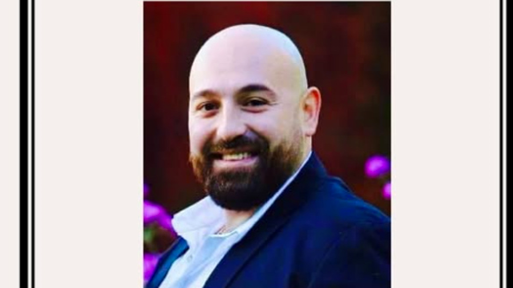 A  picture of a man with a brown beard, smiling towards the camera and wearing a white shirt and blue jacket