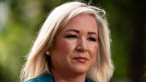 Michelle O'Neill has shoulder-length blonde hair and is wearing a turquoise suit jacket. She is standing outside. 