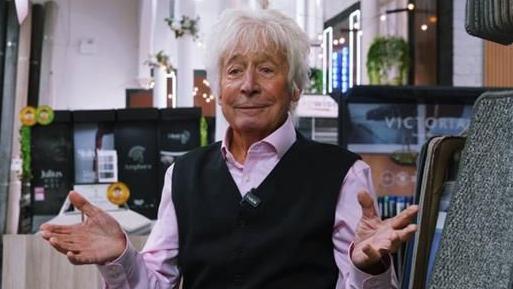 A recent photo of Mike Smith, who has long white hair. He is wearing a pink shirt with a black vest.