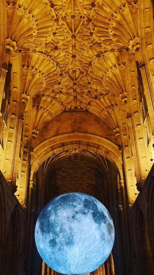 Jerram's Museum of the Moon - a giant moon artwork - lit up at the bottom of the image, surrounded by darkness, with the detailed ceiling of Sherborne Abbey above, which is also lit up.