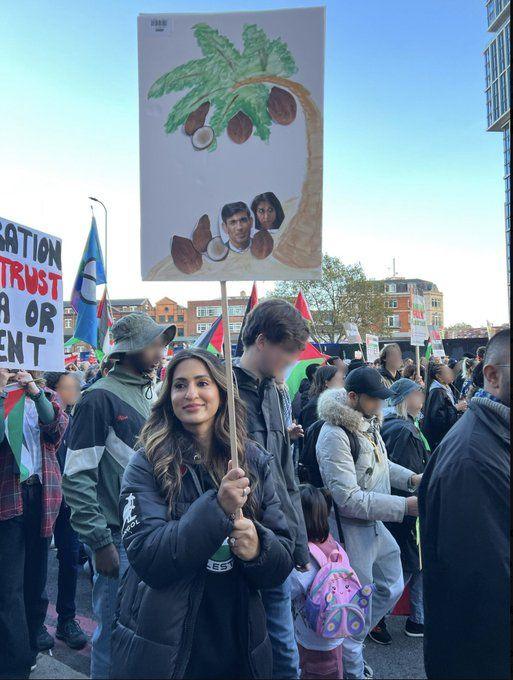 Marieha Hussain pictured on the march holding a placard, with a drawing of a palm tree with coconuts falling off it. Photos of Rishi Sunak and Suella Braverman have been stuck on top of two of the coconuts. A crowd of demonstrators can be seen in the background of the image. 