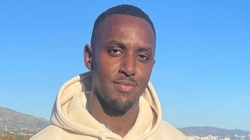 Frederick Mutala is standing in front of a large hill - to his right, the picture's left - and a sea to his left, the picture's right. He is dressed in a large white hooded top