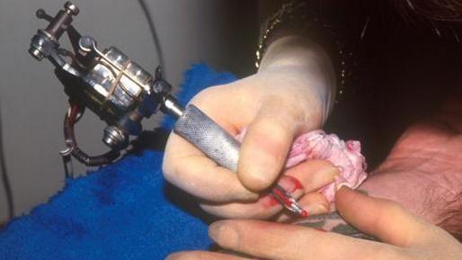 Close-up of tattoo needle being used. The Tattooist has rubber gloves on and is holding a tissue. There is some blood on the gloves.