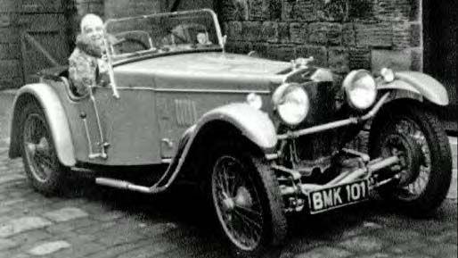 A woman sits in an old car without a roof. The number plate on the front reads BMK101.