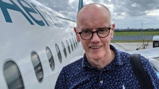 A man, John Bittles pictured beside an aeroplane. he is wearing a navy shirt, a black bag strap on his right shoulder and black glasses. He is bald.