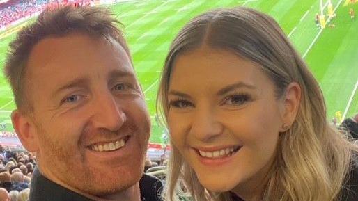 A selfie of Siobhan and her partner who are in a rugby stadium. Both are smiling at the camera from the seats with the pitch behind them. She has long wavy blonde hair and is smiling. She is in an office. He has short ginger hair and a cropped beard.