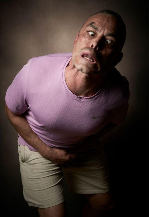 Ryan Barton has short dark hair and his tongue pushing his top lip out, and his eyes crossed. His body is also contorted with his head thrust to the side, shoulders and chest pushed forward and arms tucked up in to his waist
