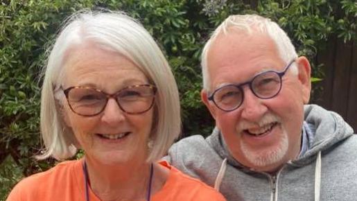 A couple pose for a picture together. Both wearing glasses and smiling, she has grey bobbed hair and wears an orange t-shirt. He has short grey hair and a goatee beard and is wearing a grey hoodie.