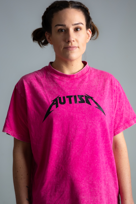 Woman with brown hair in space buns is wearing a shocking pink t-shirt with the text Autism on it. 