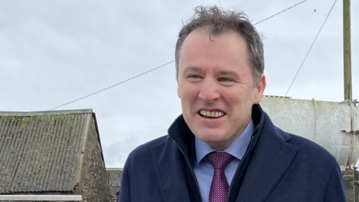 Shows a man smiling in a blue shirt, purple, navy blue overcoat and standing in front of telegraph pole with cloudy sky in the background