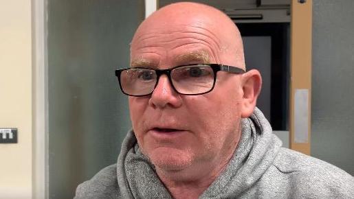 A bald man wearing black glasses and a grey hooded top stares at the camera