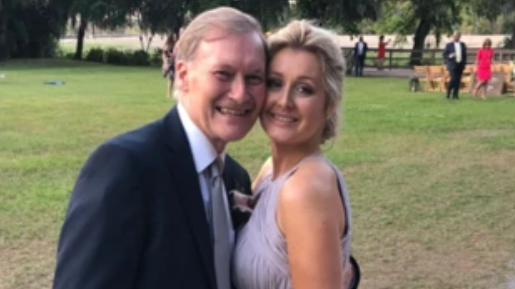 Sir David and Katie Amess standing on a grass lawn. Sir David is wearing a suit and Katie is wearing a sleeveless lilac dress. They are both smiling.