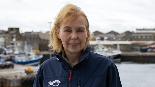 Head and shoulders of Elspeth Macdonald. She has blond hair and is wearing a blue jacket with a stylised drawing of a fish on the jacket under the right shoulder. She is standing in a small harbour which is visible in the background with a few small fishing boats there.