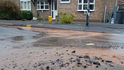 Brown water floods a residential street which has a large bump in the foreground with debris strewn across it. A pavement can be seen in the background with a house next to it.