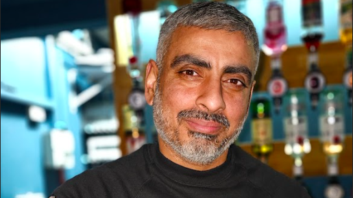 Pete is wearing a black top and is stood in the bar area of his pub