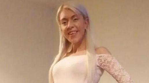 Danielle Davidson, wearing a white dress and with long blonde hair, smiling at the camera with her hands on her hips. She is in what appears to be a living room.