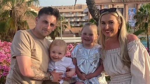 A man in a tan t-shirt, with a tattoo on his arm, and a woman in a white dress and with sunglasses on her head, stand on either side of two children - both very young - while outside what looks like a hotel.