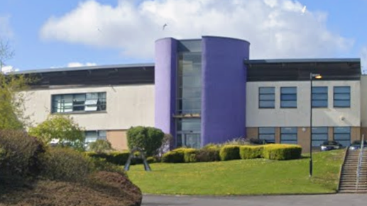 Venerable Bede Church of England Academy is a two-storey modern building, moslty in white and beige. The entrance is in its centre. It is purple and in a cylinder shape. The school is surrounded by a hedges and a grassed area with a staircase leading to the building.